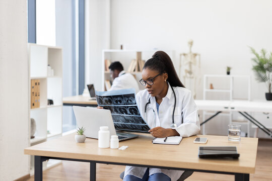Young adult female doctor examines x-ray results at desk in professional medical workspace.