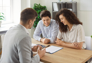 Male financial advisor consulting young couple about insurance sitting at the desk in office. Man and woman talking with realtor or business broker going to sign a contract and buy a property.