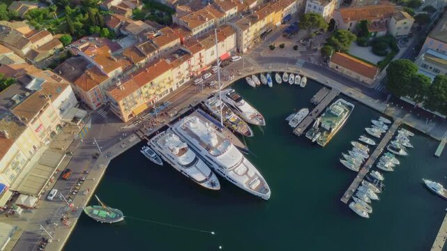 aerial drone shot capturing the vibrant Port de Saint Tropez on the French Riviera, showcasing numerous luxury yachts moored in the bustling harbor alongside picturesque waterfront buildings.