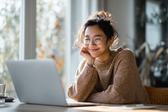 Young woman wearing glasses and a cozy sweater smiling while working on a laptop in a bright, comfortable home setting