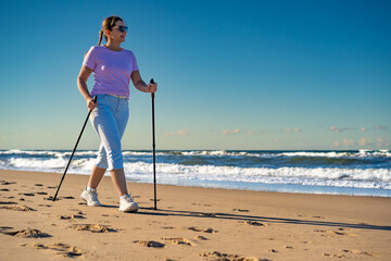 Beautiful mature woman training Nordic walking on sandy beach on summer afternoon. Side view	
