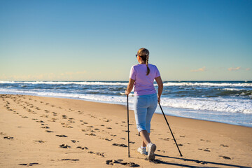 Beautiful mature woman training Nordic walking on sandy beach on summer afternoon. Back view	
