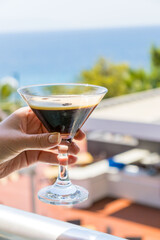 Close-up of a refreshing espresso martini cocktail in a clear martini glass, elegantly held by a woman in a white knitted summer dress. The creamy coffee foam is topped with three roasted coffee beans