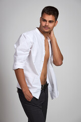 Attractive young brunette guy with short haircut in black trousers and white shirt posing in studio on white background