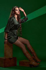 beautiful brunette girl with luxurious long hair in a green short dress, leather high boots with lacing and jewelry on her neck poses in the studio on a green background with beautiful lighting