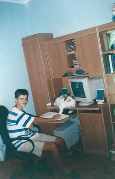 Analog photo from early 2000s Ukraine showing a teenager sitting at a home computer desk with a cat nearby, warm interior lighting