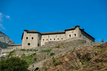 The majestic Fortress of Bard, a monumental military structure, stands imposingly on a rocky cliffside under a clear blue sky in Italy's Aosta Valley.