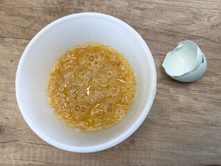 Whisked raw eggs in a white plastic bowl with broken eggshell on wooden kitchen surface, close-up detail for cooking and meal preparation.