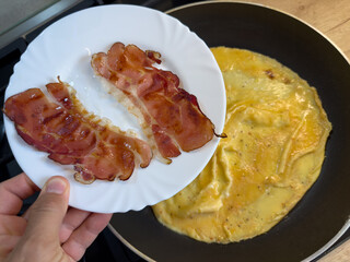 Crispy bacon slices on white plate held above frying pan with omelette cooking on kitchen stove, close-up of homemade breakfast preparation.