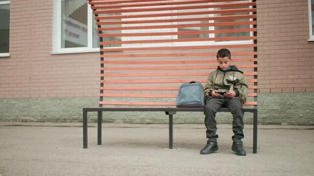 young boy sitting on bench holding phone gaming during breaktime bag beside him glances left and right before focusing back on screen calm outdoor schoolyard setting with modern bench and brick wall