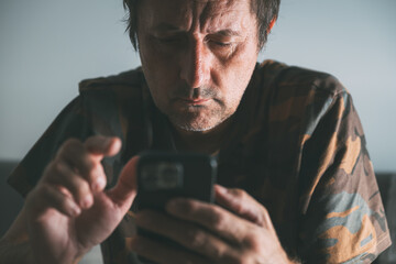 Focused middle-aged man using smartphone at home, intense concentration in low light