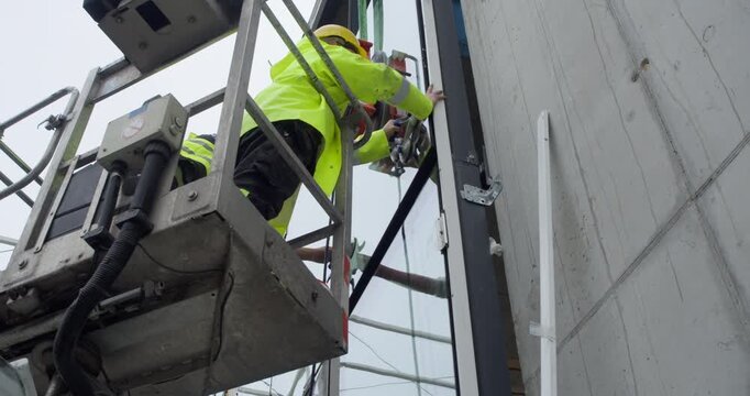 A team of glaziers uses an industrial vacuum lifter with suction cups to maneuver a heavy glass panel. The workers are guiding the large window into the frame of a modern building facade.