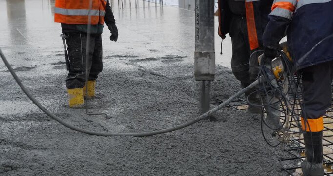 Pouring a concrete floor slab at a construction site. Wet concrete flows from a pump hose over steel rebar, while a worker uses a poker vibrator to remove air bubbles from the mixture.