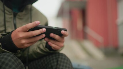 Partial view of kid in green jacket playing game on handheld device outdoors, fingers gripping phone tightly with blurred background of urban building creating casual leisure moment