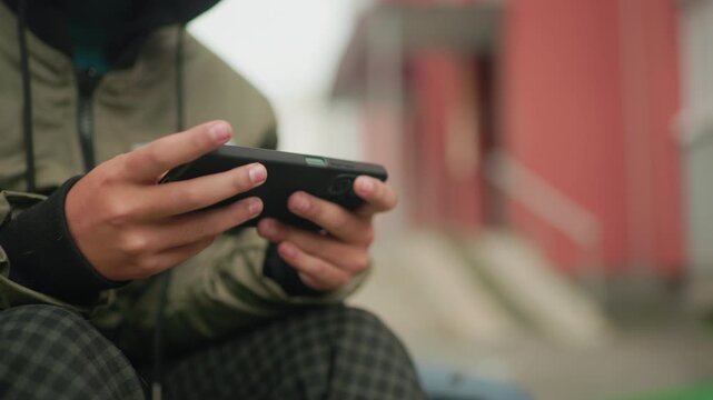 Partial view of kid in green jacket playing game on handheld device outdoors, fingers gripping phone tightly with blurred background of urban building creating casual leisure moment