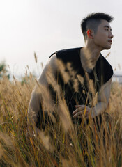 Man in a black sleeveless shirt crouching in tall grass during sunset, conveying a modern and warm...