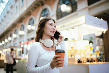 A stylish young woman, possibly an influencer, holds a coffee cup and uses her smartphone outdoors in an urban cafe environment.