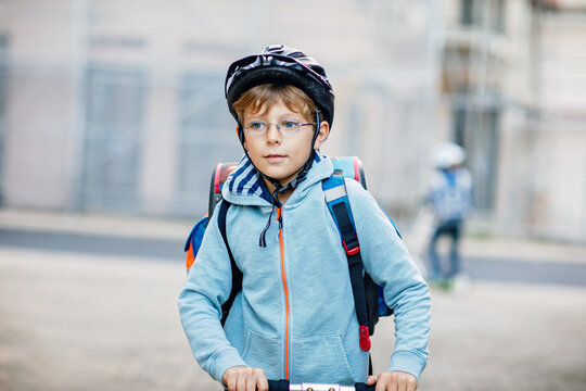 Active school kid boy in safety helmet riding with his scooter in the city with backpack on sunny day. Happy child in colorful clothes biking on way to school.