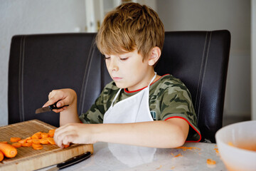 Cute school kid boy cutting fresh carrots preparing salad or snack box for school. Happy healthy child in domestic kitchen, indoors