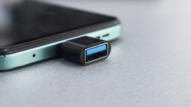 A close-up shot of a small USB OTG plugged into the charging port of a green smartphone, lying on a gray textured surface.