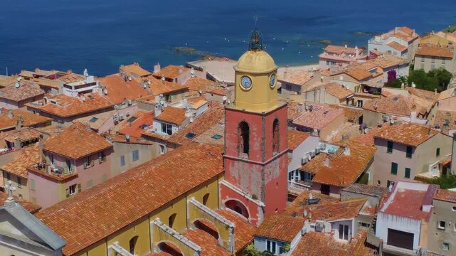 An aerial view showcases the iconic orange tiled rooftops of Saint Tropez, France, with the prominent yellow and red bell tower of the Church of Notre Dame de l'Assomption dominating the charming vill