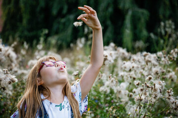 Fototapeta premium Portrait of happy elementary school girl with glasses playing outdoors in summer meadow, blowing fluffy seeds in nature. Real life authentic childhood moment.