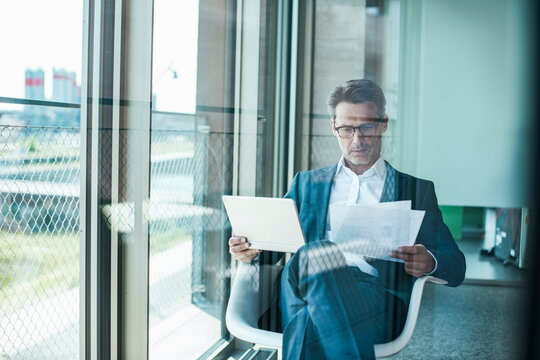 Businessperson seated in modern office reading paperwork and using tablet