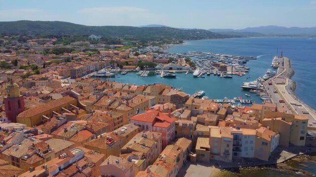 aerial drone shot capturing the picturesque harbor of Saint Tropez, France, bustling with yachts and boats, alongside the charming red-tiled rooftops of the historic old town and its iconic bell tower