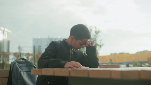 Annoyed kid sitting outdoors slams pen on wooden table in frustration, expressing irritation and stress during study session, with backpack and book beside him in urban environment
