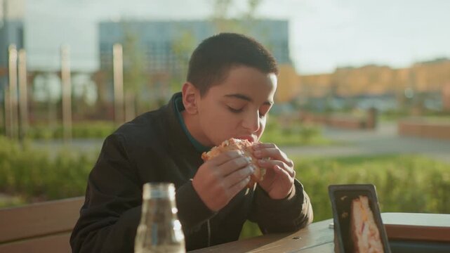 Happy boy sitting outdoors enjoying bite of pizza with juice bottle on wooden table, savoring meal with content expression under warm sunlight in casual park setting