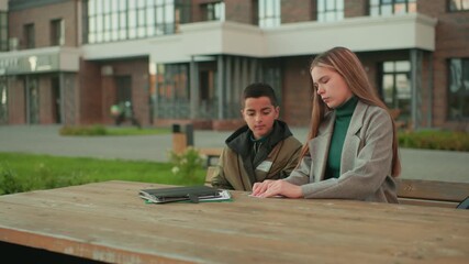 Little kid nods head while sitting beside aunt folding paper at wooden table outdoors, both engaged in learning activity with notebooks in background and urban setting around them - Powered by Adobe