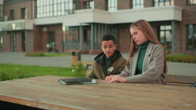 Little kid nods head while sitting beside aunt folding paper at wooden table outdoors, both engaged in learning activity with notebooks in background and urban setting around them