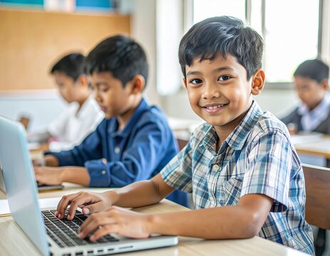 Asian boy typing on a laptop in a modest classroom, smiling.