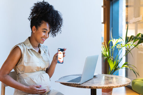 Smiling black pregnant woman showing baby ultrasound picture to laptop
