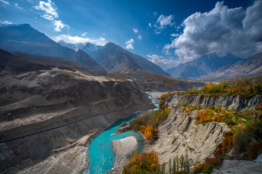 View of turquoise river snaking through rugged mountains and autumnal trees under a partly cloudy sky, Hunza Nagar, Gilgit Baltistan, Pakistan.
