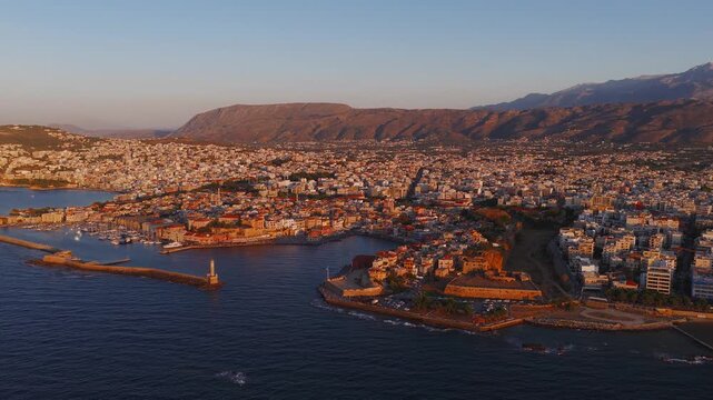 Aerial view of Chania town in Crete, Greece, during sunset. The video pans over white and pastel buildings, the harbor, and Lefka Ori mountains.