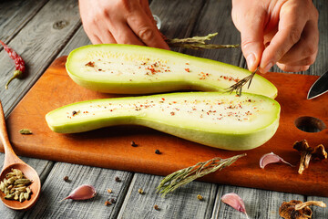 Hands prepare fresh green squash by slicing it open, revealing its soft interior. Sprigs of herbs...
