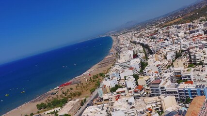 Aerial view of Rethymno, Crete, showing a sandy beach with sunbeds, a vibrant promenade, whitewashed buildings, and boats on the deep blue sea. - Powered by Adobe