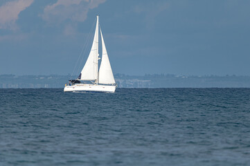 Fototapeta premium Sailboat sailing on blue water with distant coastline under cloudy sky