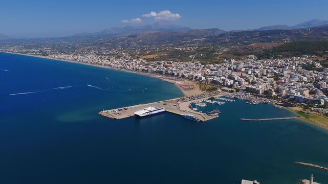 Aerial view of Rethymno, Crete, showing a harbor with ferries and yachts, a sandy beach, urban buildings, rolling hills, and a calm sea under a clear sky.