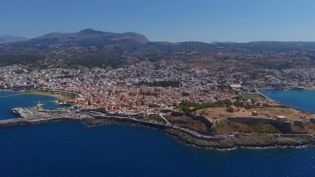Aerial view of Rethymno, Crete showing the lighthouse, curved breakwater, moving boats, white coastal buildings, and mountainous backdrop.