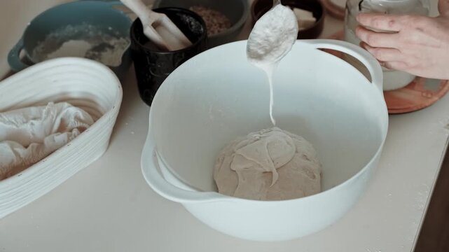 Close-up video of hands spooning live sourdough starter from a jar into bread dough in a white bowl, beginning fermentation.