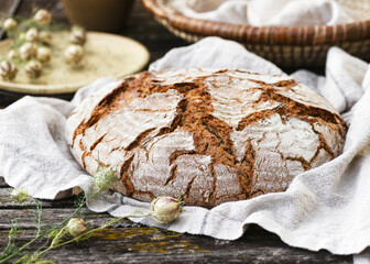 Golden, crusty bread lies on a soft cloth, its rugged surface full of character. Rustic details with wood, wicker and blossoms create a cozy farmhouse atmosphere. Healthy handmade food concept.