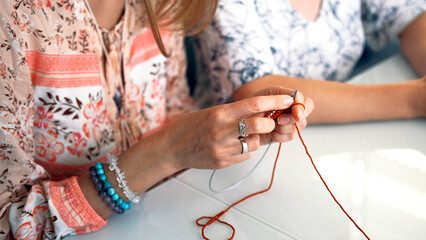 A woman knits a scarf made of red yarn, a close-up of her hands. Her friend is sitting next to her. Creativity, hobbies, and knitting