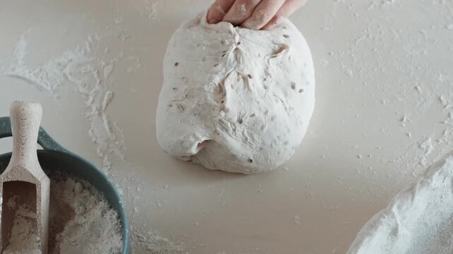 Close-up video of hands sealing and rolling bread dough, dusting a banneton with flour, placing the oval loaf inside, covering with towel.