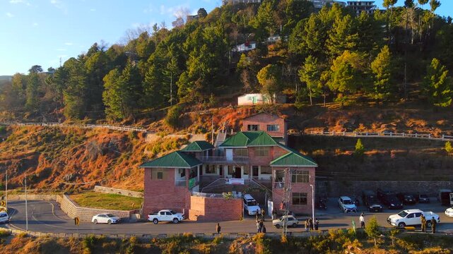 Aerial view of a building with green roofs nestled on a hill with winding roads, amidst a mix of lush green trees and warm, earthy tones, Murree, Punjab, Pakistan.