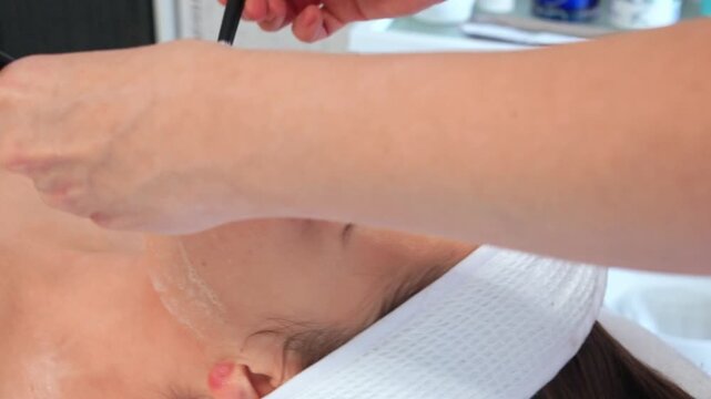 Closeup of skilled beautician applying hydrating medical peeling cream mask to young woman face, focusing on forehead. Pampering spa dermapen procedure for youthful skin care