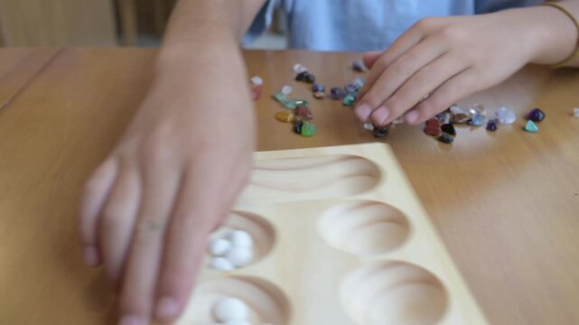 Close-up of child s hands playing mancala Kalah board game with stones. Traditional wooden board game, strategy and focus