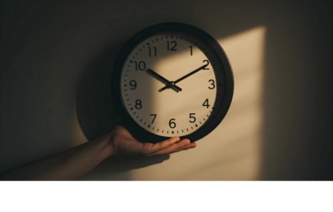 A person's hand holding a simple black and white analog clock in a beam of light, symbolizing the precious and fleeting nature of time