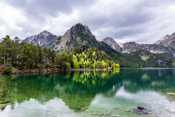 Clear alpine lake reflecting forest and snowy peaks under dramatic cloudy sky. Serene mountain lake with forest reflections and cloudy sky in peaceful nature landscape. Crystal-clear alpine lake.